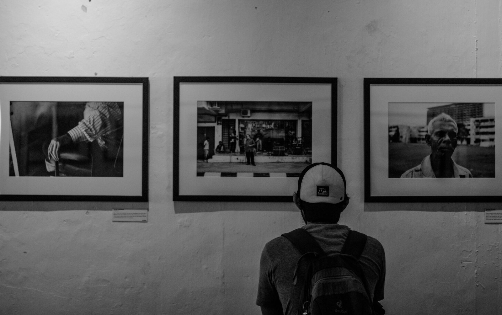 A man in a cap and backpack admires monochrome art in a gallery.